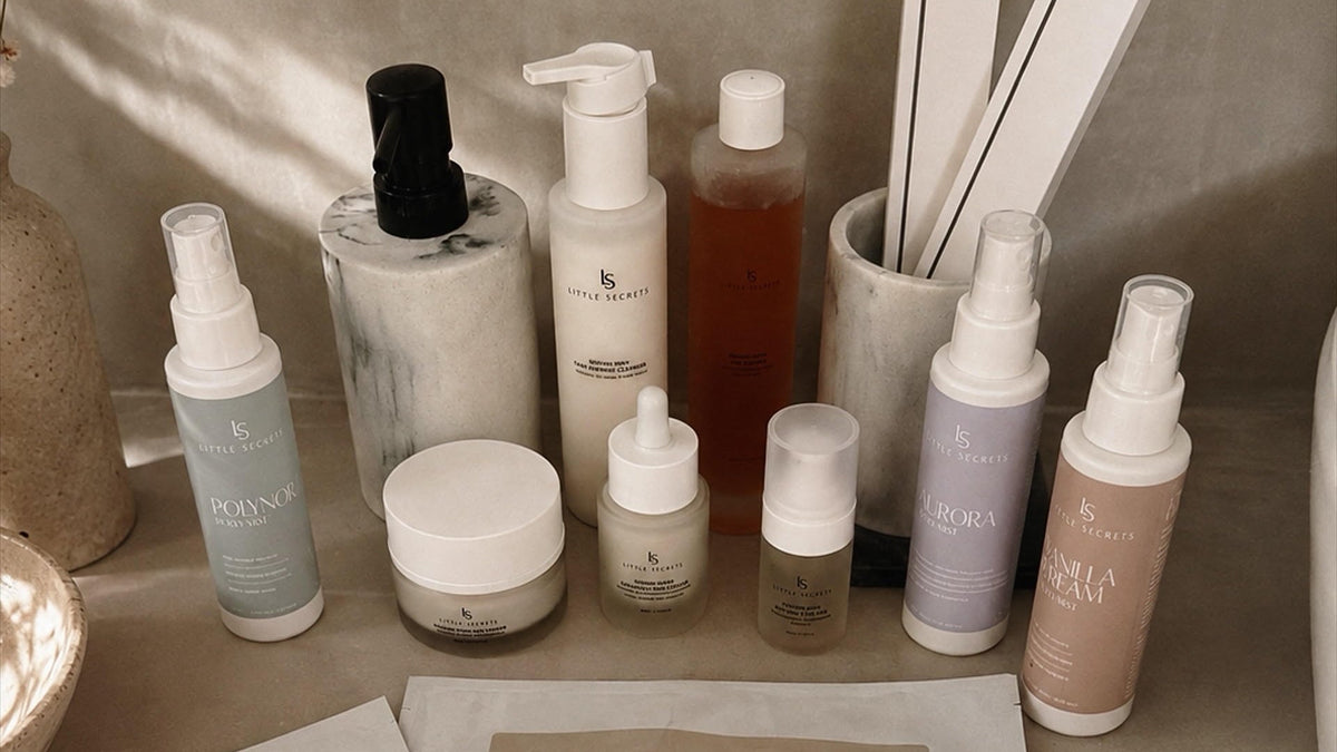 A collection of Little Secrets skincare products arranged on a beige stone surface, including several facial mask packets in the foreground and multiple skincare bottles and jars behind them.