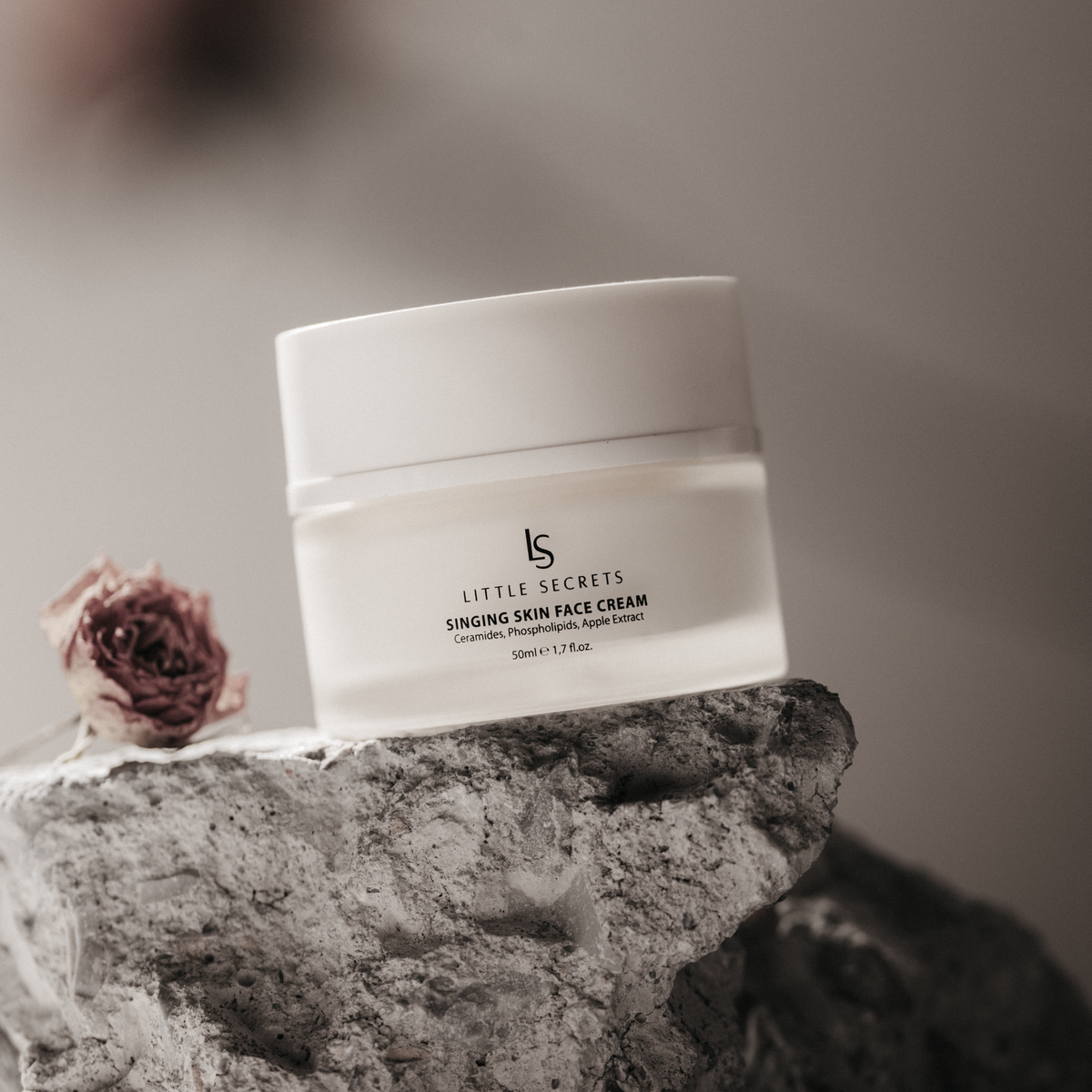 A white jar of Little Secrets Singing Skin Face Cream styled on a textured gray stone with dried rose petals, set against a soft, warm neutral background.