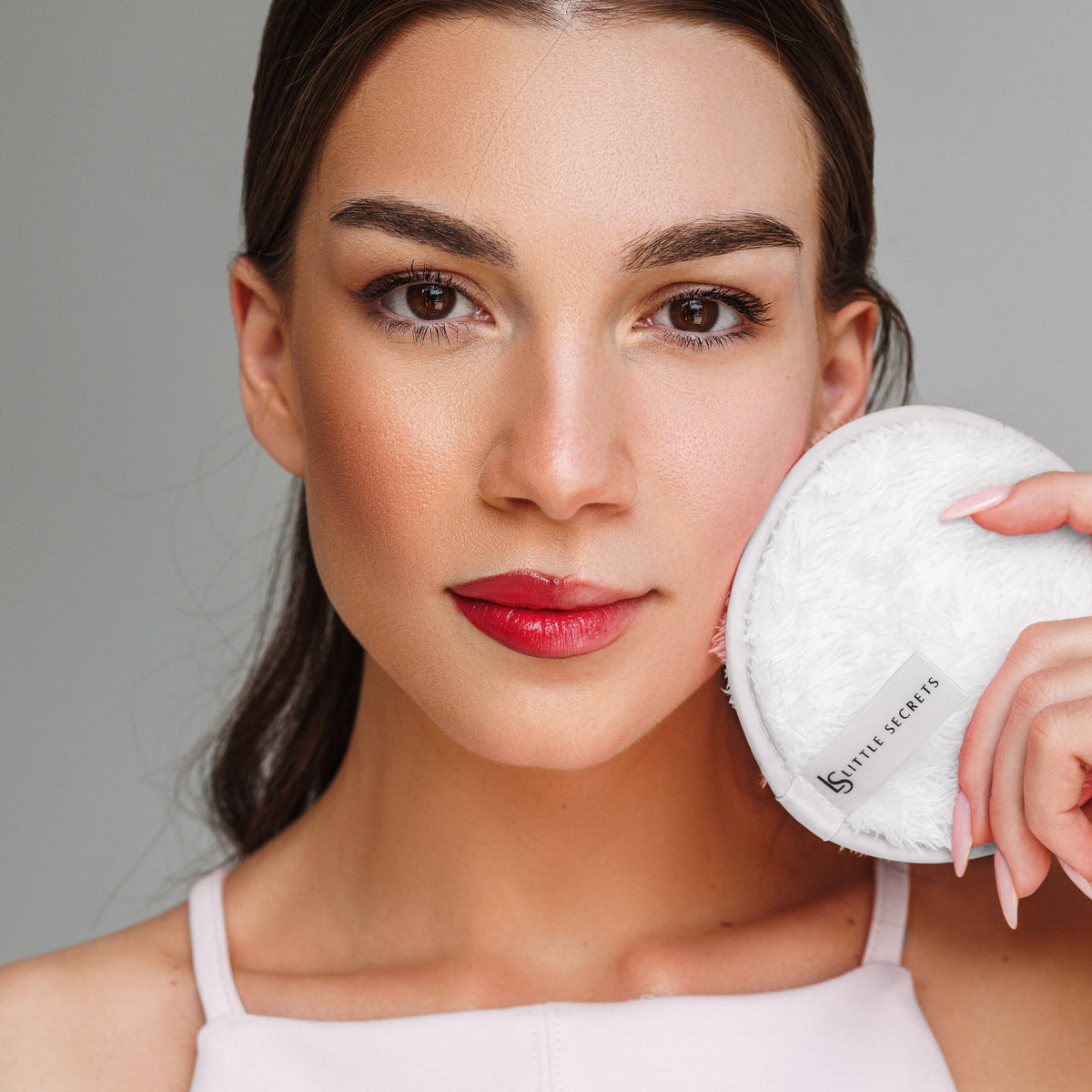 A close-up of a woman holding a round white makeup puff next to her face, wearing a light pink top against a solid gray background. The makeup puff has a visible label reading 'little secrets'.