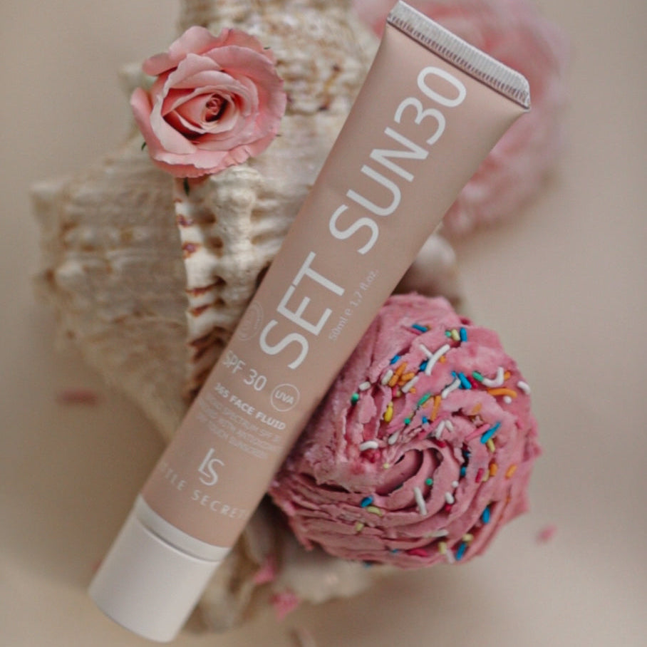A tube of SET SUN30 SPF 30 sunscreen is styled diagonally on a beige background, surrounded by decorative pink roses and a seashell.