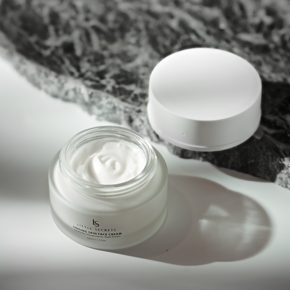 A glass jar of Little Secrets Hyalu-Firming Cream with white cream visible inside, shown next to its white lid on a white surface with a dark gray marble prop in the background.
