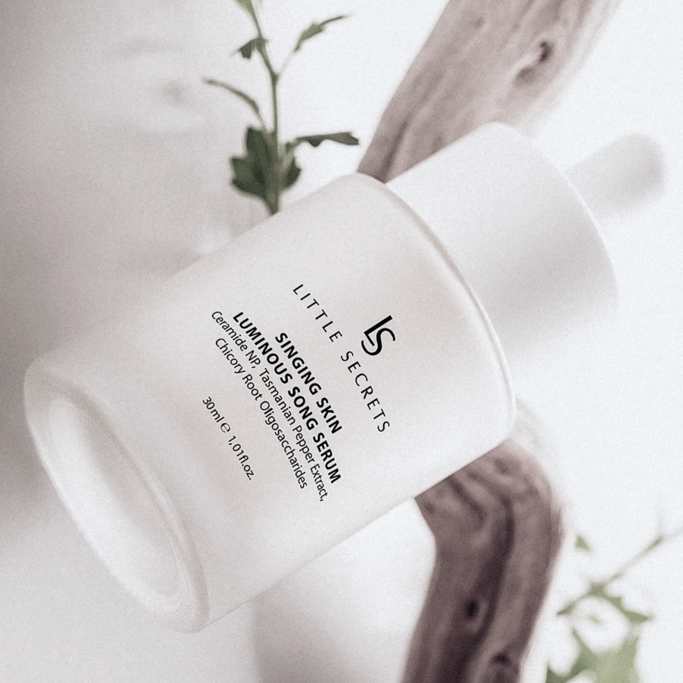 A white bottle of Little Secrets Singing Skin Luminary Sun Serum, shown at an angle on a light background with a piece of driftwood and green leaves.