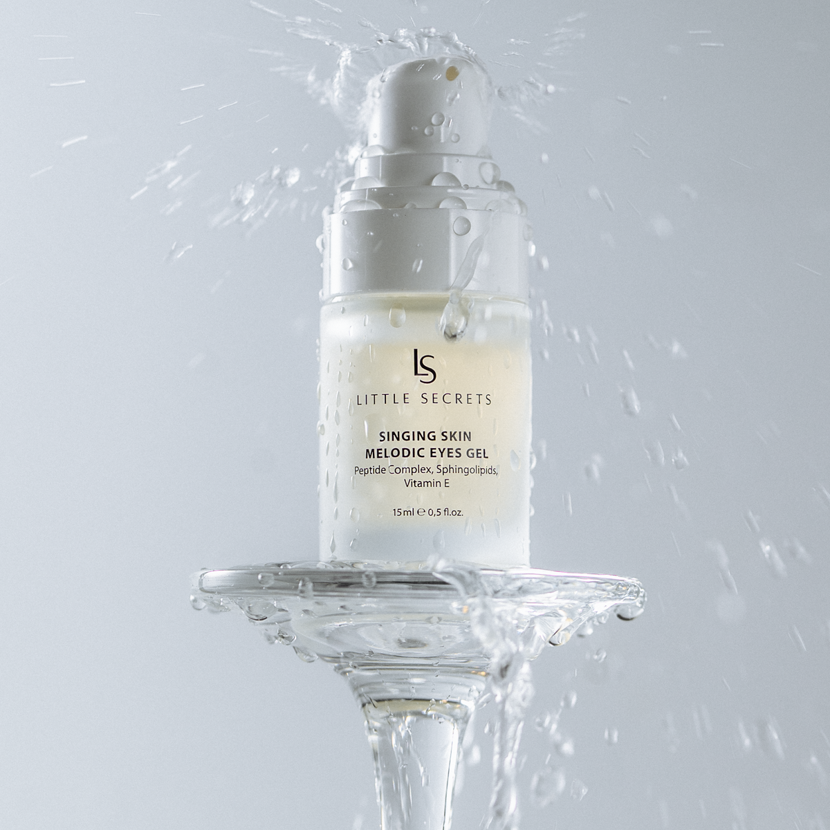 A bottle of Little Secrets Singing Skin Melinose Eye Oil suspended above a clear glass pedestal, surrounded by dramatic water droplets and splashes against a light gray background.