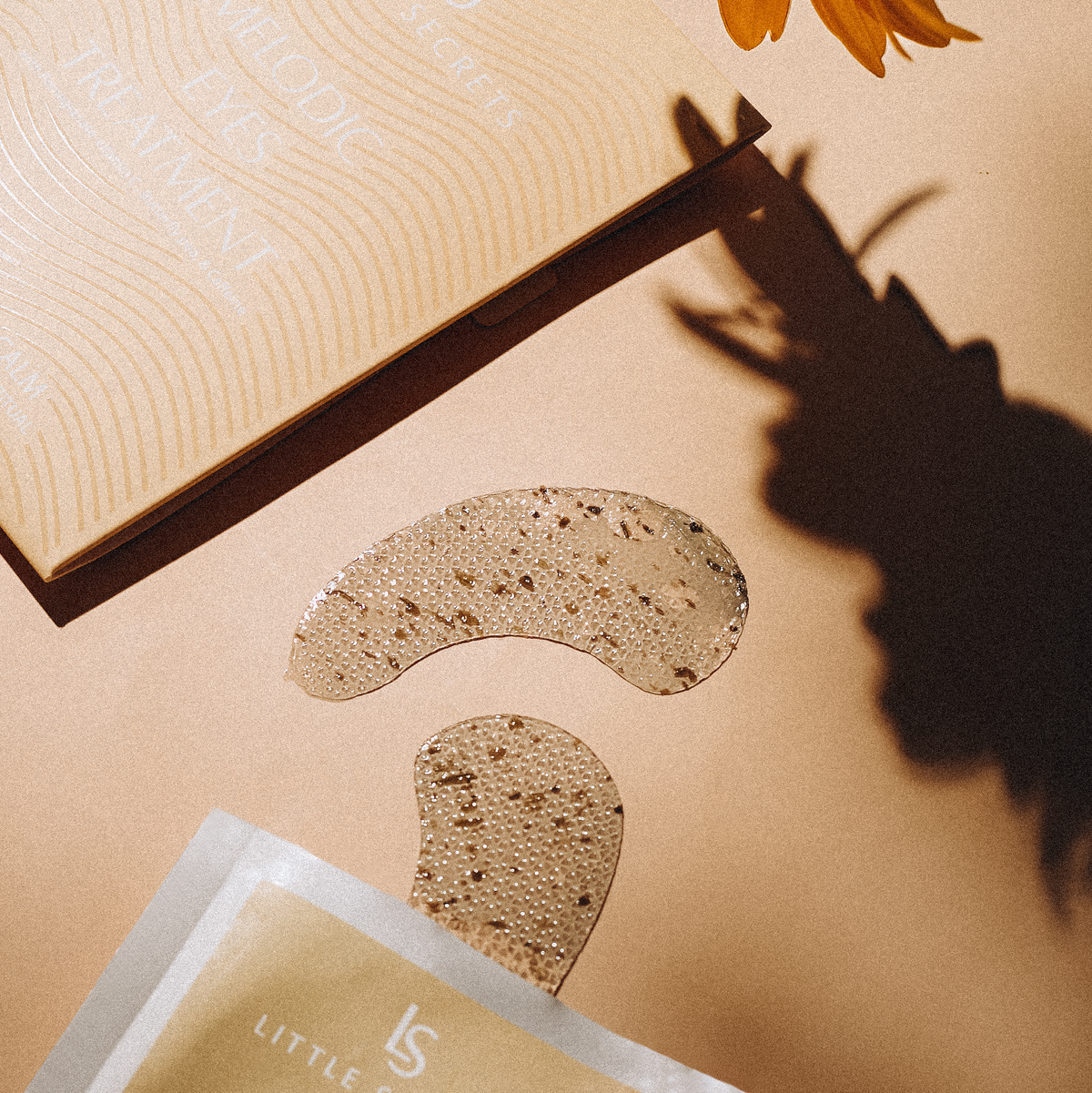 Two beige patchouli gel eye masks are displayed on a warm beige background next to their packaging and a yellow flower, with a prominent shadow cast on the right.