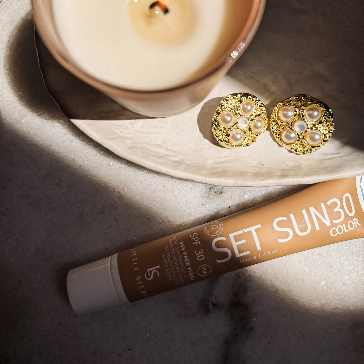 A tube of Little Sister Set Sun 30 Color SPF 30 sunscreen lies on a gray marble surface next to a lit candle and a pair of gold pearl stud earrings.