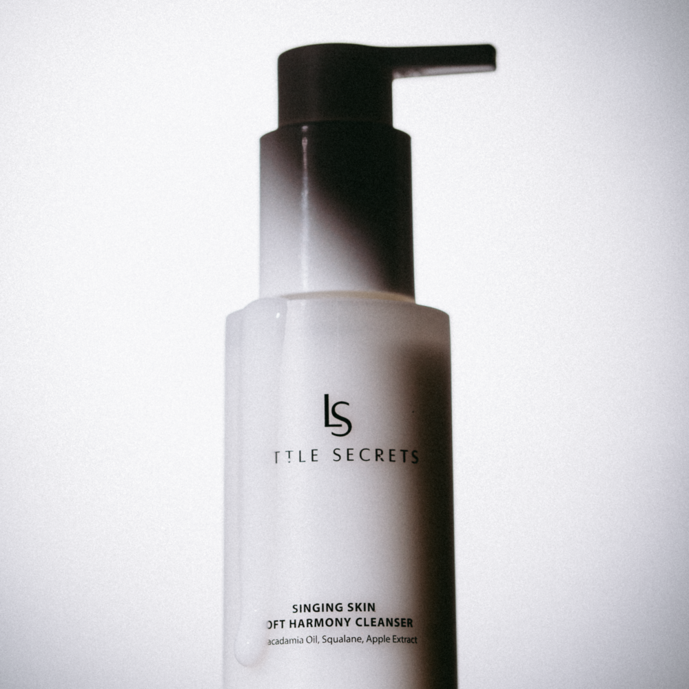 A bottle of Little Secrets Singing Skin Jet Harmony Cleanser with a frosted white body and a black pump dispenser, set against a plain light gray background.