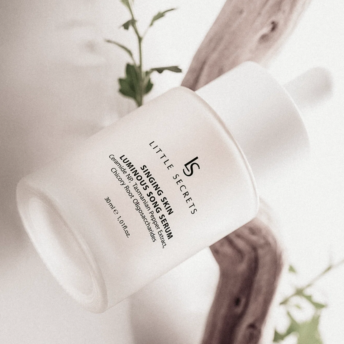 A frosted white bottle of Little Secrets Singing Skin Serum with a dropper cap, styled diagonally on a light background with a piece of driftwood and green leaves.